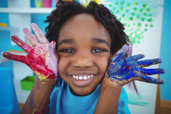 A child with a large smile hold hands covered in blue and red paint up to their face